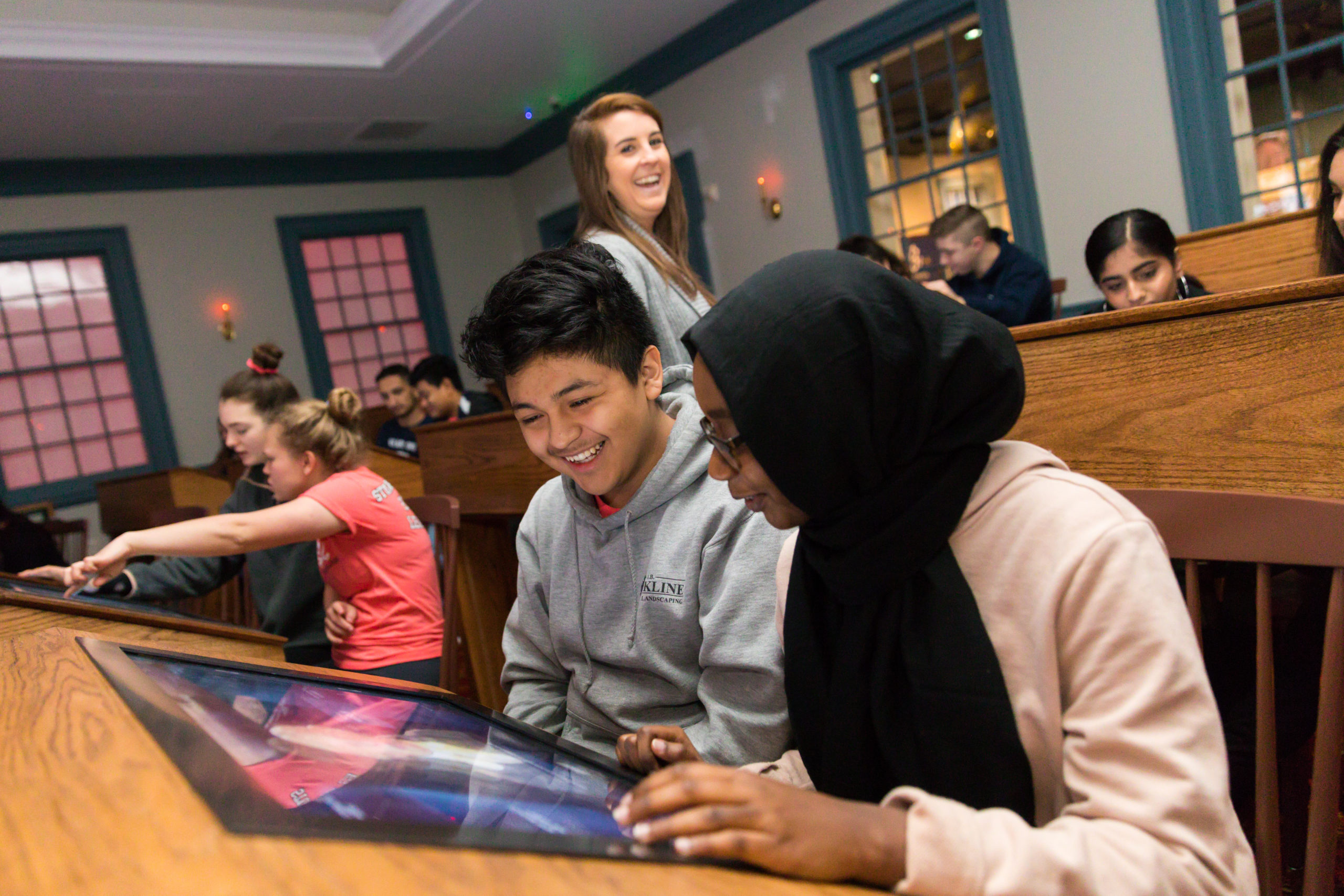 Two students interact while looking at a screen at a desk. A teacher and other students are behind them.