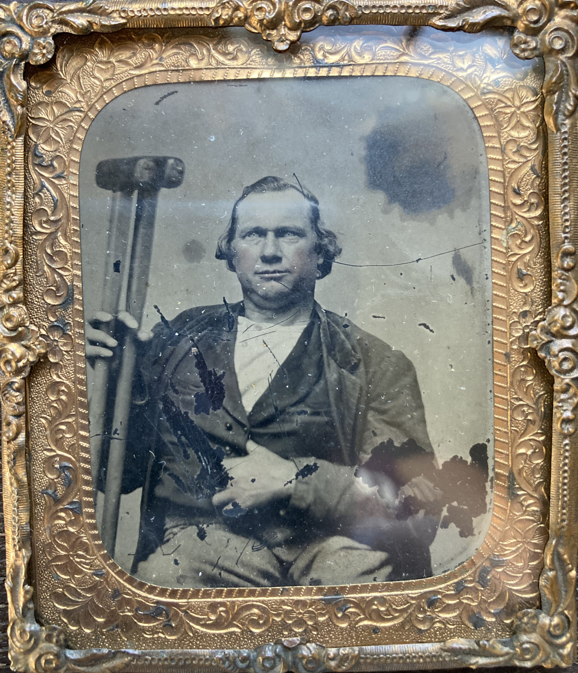 1860s tintype of a man in a gold frame. Man is seated, holding two crutches in his right hand. Left hand tucked into coat. Cheeks tinted pink.